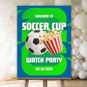以下が含まれることがあります： イーゼルに立てられた看板で、白い文字で「Welcome to Soccer Cup Watch Party」と書かれています。看板にはサッカーボール、ポップコーンのバケツ、日付06.20.2026が描かれています。背景は緑と青です。