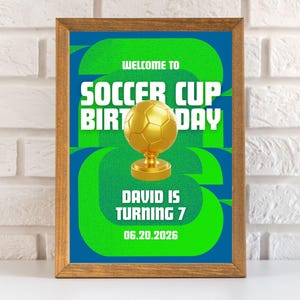 以下が含まれることがあります： サッカーをテーマにした誕生日のパーティーのサインが額縁に入っています。 金色のサッカーボールのトロフィーと、緑と白で「Welcome to Soccer Cup Birthday Day. David is turning 7, 06.20.2026」と書かれたテキストが、青と緑の背景にあります。