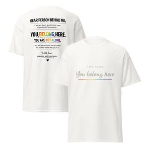 May include: Two white t-shirts with text. One shirt says "DEAR PERSON BEHIND ME, YOU BELONG HERE. YOU ARE NOT ALONE." The other shirt says "SAFE SPACE You belong here" with a rainbow underline. The shirts are a casual style.
