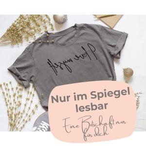 Può includere: Una maglietta grigia con la scritta "Azum and P" in stile manoscritto. La maglietta ha le maniche arrotolate. Una grafica ovale rosa in basso recita "Nur im Spiegel lesbar. Eine Botschaft nur für dich."