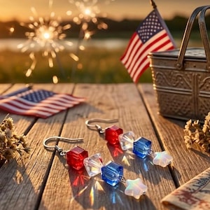May include: A pair of patriotic earrings with red, white, and blue beads and star charms. The earrings are displayed on a wooden surface with an American flag, a picnic basket, and sparklers in the background.