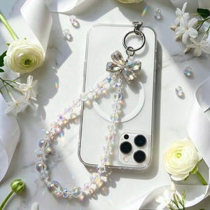 May include: A clear smartphone case with a circular magnetic ring on the back, attached to a long beaded strap. The strap features iridescent, translucent spherical beads and a silver-toned butterfly charm with clear crystals at the top.