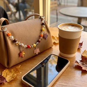 May include: A tan textured handbag featuring a decorative beaded strap with colorful autumn-themed charms including orange, yellow, and brown beads interspersed with clear crystals and small maple leaf pendants in shades of red and brown.