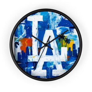May include: Circular wall clock with a black frame. The clock face displays a blue and white abstract design with the letters "LA" in white. Palm trees and a cityscape are also visible.