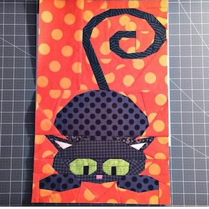 Può includere: Un gatto nero con la coda arricciata, occhi verdi e naso rosa. Il gatto è fatto di stoffa e si trova su uno sfondo rosso con pois arancioni.