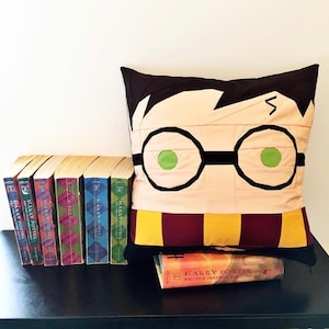 Pode incluir: Uma almofada com tema Harry Potter com um design de tecido preto e castanho. A almofada apresenta uma imagem de desenho animado de Harry Potter com os seus óculos e a sua cicatriz em forma de raio. A almofada está sentada sobre uma superfície preta com uma cópia do livro "Harry Potter e as Relíquias da Morte" à sua frente.