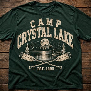 Puede incluir: Una camiseta de manga corta de color verde oscuro con un estampado gráfico desgastado que dice CAMP CRYSTAL LAKE en la parte superior y EST. 1980 en la inferior. El diseño incluye luna llena, montañas, pinos, un lago y dos remos cruzados.