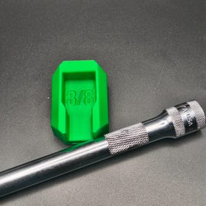 May include: A bright green plastic socket holder with the text 3/8 embossed on the center, positioned next to a silver metal socket extension bar with a knurled grip and the text USA engraved on the side of the cylindrical shaft.