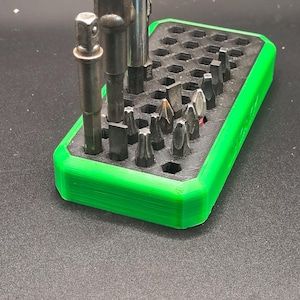 May include: A black and green 3D-printed tool organizer holding various metal bits and sockets. The organizer has a rectangular shape with a bright green border and a honeycomb pattern of holes to hold the tools. The tools are of different sizes and shapes.