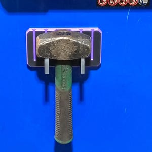 May include: A black and purple rectangular magnetic tool holder mounted on a blue surface, securely holding a heavy-duty hammer with a dark green handle and a metallic head. The holder features vertical slots and a clean, functional design.