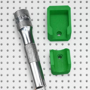 May include: A silver metal tool extension bar with a knurled grip and the text CX5KA, accompanied by two green 3D-printed plastic mounting brackets, each featuring a central circular hole for attachment purposes on a white pegboard surface.