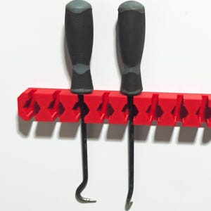 May include: A red tool holder with two black and gray handled tools. The holder is mounted on a white wall. The tools are hook and pick tools.