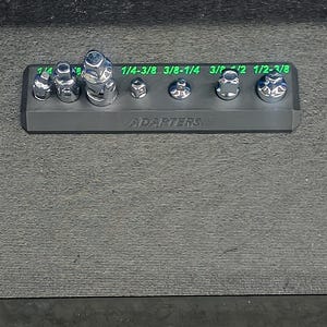 May include: A set of chrome adapters of varying sizes, arranged on a black holder. The sizes are labeled in green text: 1/4-3/8, 3/8-1/4, 3/8, 1/2, and 1/2-3/8. The word "ADAPTERS" is printed on the holder.
