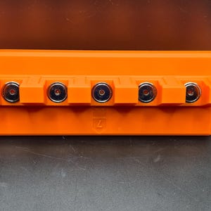 May include: An orange rectangular object with five silver-colored circular components. The object has a geometric design and is set against a dark background. The number "7" is visible on the object.