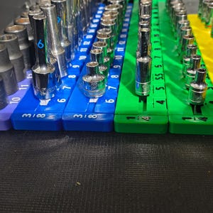 May include: A collection of metal socket tools organized on multi-colored plastic rails. The set includes various sizes of sockets arranged on purple, blue, green, and yellow holders, with numerical markings printed on the base of each rail.
