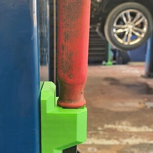 May include: A bright green 3D-printed plastic tool holder mounted to a blue metal surface. The holder secures a metal tool with a worn, cylindrical red rubber handle that extends vertically from the top of the green bracket.