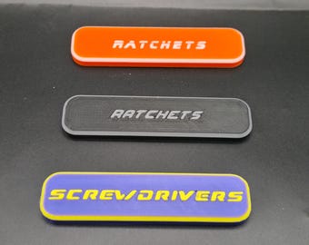 May include: Three rectangular labels with rounded corners. The top label is orange with white text reading RATCHETS. The middle label is black with grey text reading RATCHETS. The bottom label is purple with yellow text reading SCREWDRIVERS.