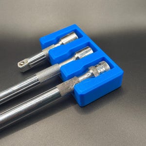 May include: A blue 3D-printed tool organiser holding three silver socket wrenches. The wrenches are arranged in a row, with their textured handles and socket ends visible. The organiser is on a dark surface.