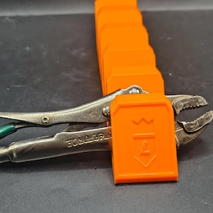 May include: A silver metal locking plier with a green handle grip is held in place by a bright orange plastic organizer rack. The rack features a series of vertical slots, with the front slot embossed with a crown icon and the number 7.