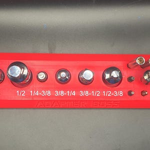 May include: A red adapter holder with various sized sockets and bits. The holder has the text "ADAPTER BOSS" and measurements like 1/4, 3/8, 1/2, 1/4-3/8, 3/8-1/4, 3/8-1/2, and 1/2-3/8.