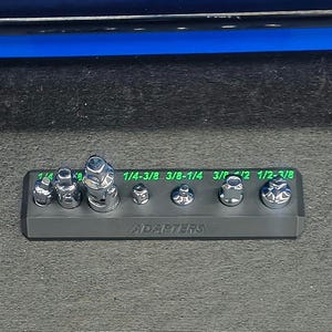 May include: A black plastic socket adapter organizer tray holding seven chrome-plated steel adapters of various sizes. The tray features bright green printed text indicating sizes such as 1/4-3/8 and 3/8-1/4, with the word ADAPTERS embossed on the front base.