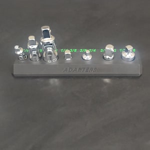 May include: A set of chrome-plated socket adapters of various sizes, arranged on a dark gray holder. The sizes are labeled in green. The word "ADAPTERS" is printed on the holder.