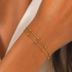 May include: A delicate double-strand gold bracelet featuring small, evenly spaced polished gold beads along each thin chain. The two chains are connected, creating a layered appearance with a bright, reflective metallic finish throughout.