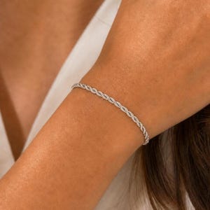 May include: A silver rope chain bracelet. The bracelet is worn on an arm. The bracelet has a twisted metal design. The bracelet is a simple, elegant piece of jewellery.