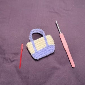 May include: A miniature crochet bag with a cream-colored body and lavender trim and handles. A red needle and a pink crochet hook are also present on a purple surface. The bag is a handmade item.
