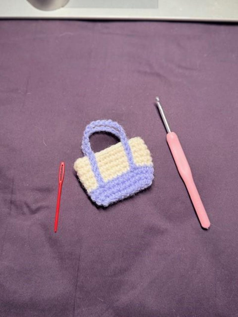 May include: A miniature crochet bag with a cream-colored body and lavender trim and handles. A red needle and a pink crochet hook are also present on a purple surface. The bag is a handmade item.