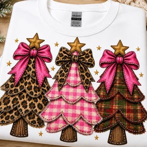 May include: A white crewneck sweatshirt featuring a graphic of three stylized Christmas trees. The left tree has a leopard print, the middle tree has a pink gingham pattern, and the right tree has a red and green plaid pattern, all topped with gold stars and bows.