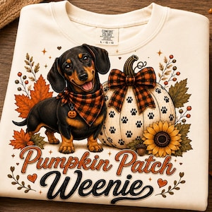 May include: A cream-colored t-shirt featuring a graphic of a black and tan dachshund wearing a plaid bandana next to a white pumpkin with paw prints, a plaid bow, and a sunflower. The text reads Pumpkin Patch Weenie in brown cursive script.