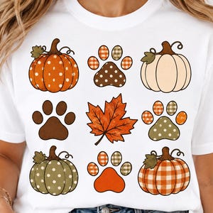 Puede incluir: Una camiseta blanca de manga corta con una cuadrícula de nueve gráficos de temática otoñal. El diseño incluye calabazas con patrones de lunares y cuadros, varias huellas de patas con detalles de lunares y cuadros, y una hoja de arce.