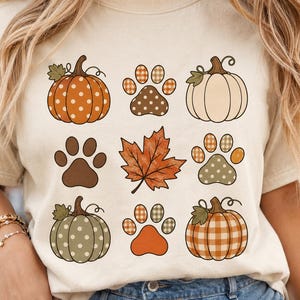 Puede incluir: Una camiseta de color crema con un gráfico de cuadrícula de iconos con temática otoñal. El diseño incluye calabazas con patrones de lunares y cuadros, varias huellas de animales con detalles a cuadros y puntos, y una hoja de arce naranja.