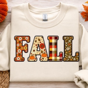 May include: A cream-colored crewneck sweatshirt featuring the word FALL printed in large block letters. Each letter has a unique pattern: the F has orange flowers, the A has pumpkins and flowers, the L has a red and yellow plaid, and the L has green leaves.