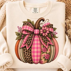 May include: A cream-coloured crewneck sweatshirt featuring a graphic of a pumpkin decorated with pink gingham, leopard print, and floral patterns. A large pink gingham and leopard print bow sits atop the pumpkin, accented by pink flowers and berries.