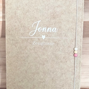 Könnte beinhalten: Ein beigefarbener Filzordner mit einem weißen Kordelverschluss und drei bunten Perlen. Der Ordner trägt den Namen "Jonna" und das Wort "Zeugnisse" in weiß.
