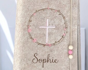 Könnte beinhalten: Ein beigefarbener Filzbuchumschlag mit einem weißen Kreuz in einem rosafarbenen Blumengekränz. Der Name "Sophie" ist unter dem Kranz gestickt. Ein weißes Band mit rosa Perlen ist am Umschlag befestigt.