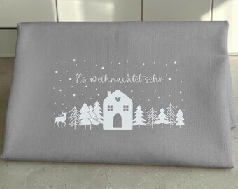 May include: A gray fabric piece featuring a white graphic print of a house, pine trees, a deer, and stars. The text "Es weihnachtet sehr" is printed in white script above the house. The design includes a snowy scene with falling stars.