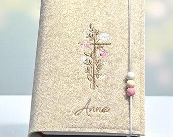 Könnte beinhalten: Beige Filz-Buchhülle mit gesticktem Kreuzdesign mit rosa und weißen Blumen. Der Name "Anna" ist darunter gestickt. Ein weißes Gummiband mit drei dekorativen Perlen sichert die Hülle.