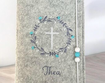 Könnte beinhalten: Grauer Filzbuchumschlag mit einem weißen Kreuz in einem blauen Blumenkranz. Der Name Thea ist unter dem Kranz gestickt. Der Umschlag hat eine weiße Schnur mit blauen Perlen.