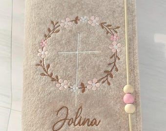 Könnte beinhalten: Ein beigefarbener Filzbuchumschlag mit einem weißen Kreuz, das in einen rosafarbenen Blumengirlanden gestickt ist. Der Name "Jolena" ist unter dem Kranz gestickt. Der Umschlag hat einen braunen Riemen mit rosa und braunen Holzperlen.