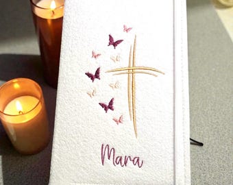 Könnte beinhalten: Weißer Bucheinband mit gesticktem goldenen Kreuz und Schmetterlingen. Der Name "Mara" ist in Burgunderrot gestickt. Zwei brennende Kerzen sind im Hintergrund.
