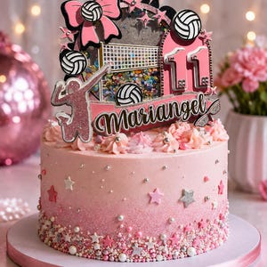 Puede incluir: Un pastel rosa degradado decorado con chispas de estrellas plateadas y rosas, coronado con un adorno de voleibol con el nombre Mariangel, el número 11, balones, una red, un lazo rosa y la silueta de una persona jugando voleibol.