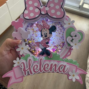 Könnte beinhalten: Rosa-weiße Minnie-Maus-Tortenaufsatz mit dem Namen "Helena" auf einem Banner. Der Aufsatz zeigt eine große Schleife mit Tupfen, eine Minnie-Maus-Figur und die Zahl "3" in Herzform, umgeben von Blumen.