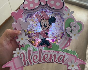 May include: A pink and white Minnie Mouse-themed cake topper with the name "Helena" on a banner. The topper features a large polka-dot bow, a Minnie Mouse figure, and a number "3" in a heart shape, surrounded by flowers.