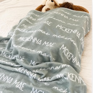 May include: A soft, light blue blanket with the name "MCKENNA MAE" in white script repeated across the surface. A stuffed animal and a child's head are partially visible under the blanket. The blanket is folded on a bed.