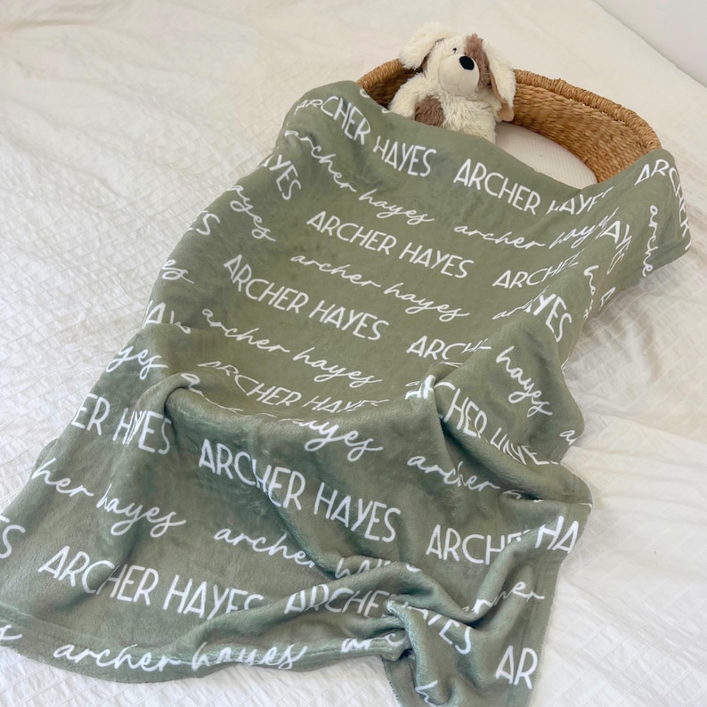 Puede incluir: Una manta de color verde salvia con el texto blanco repetido Archer Hayes en fuentes de bloque y cursiva. Un peque&ntilde;o peluche de perro marr&oacute;n claro y blanco est&aacute; metido en el borde superior de la manta, drapeada sobre una cesta tejida.