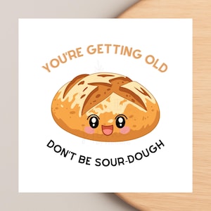 May include: A square greeting card featuring a cartoon sourdough loaf with a smiling face, pink cheeks, and large black eyes. The text above reads YOU'RE GETTING OLD and the text below reads DON'T BE SOUR-DOUGH in black capital letters.