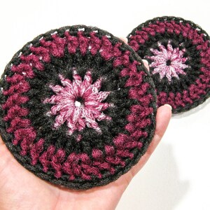 May include: Two handmade crochet coasters. Each coaster features a central starburst design in pink and white, surrounded by a ring of dark gray, and a border of deep burgundy. The coasters are round and appear to be made of yarn.